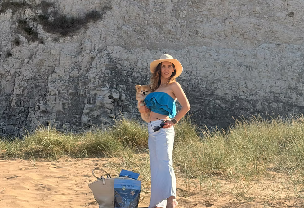 A Dr Veneta wearing a straw hat, a blue strapless tube top, and a long white skirt stands on a sandy beach holding a small dog.