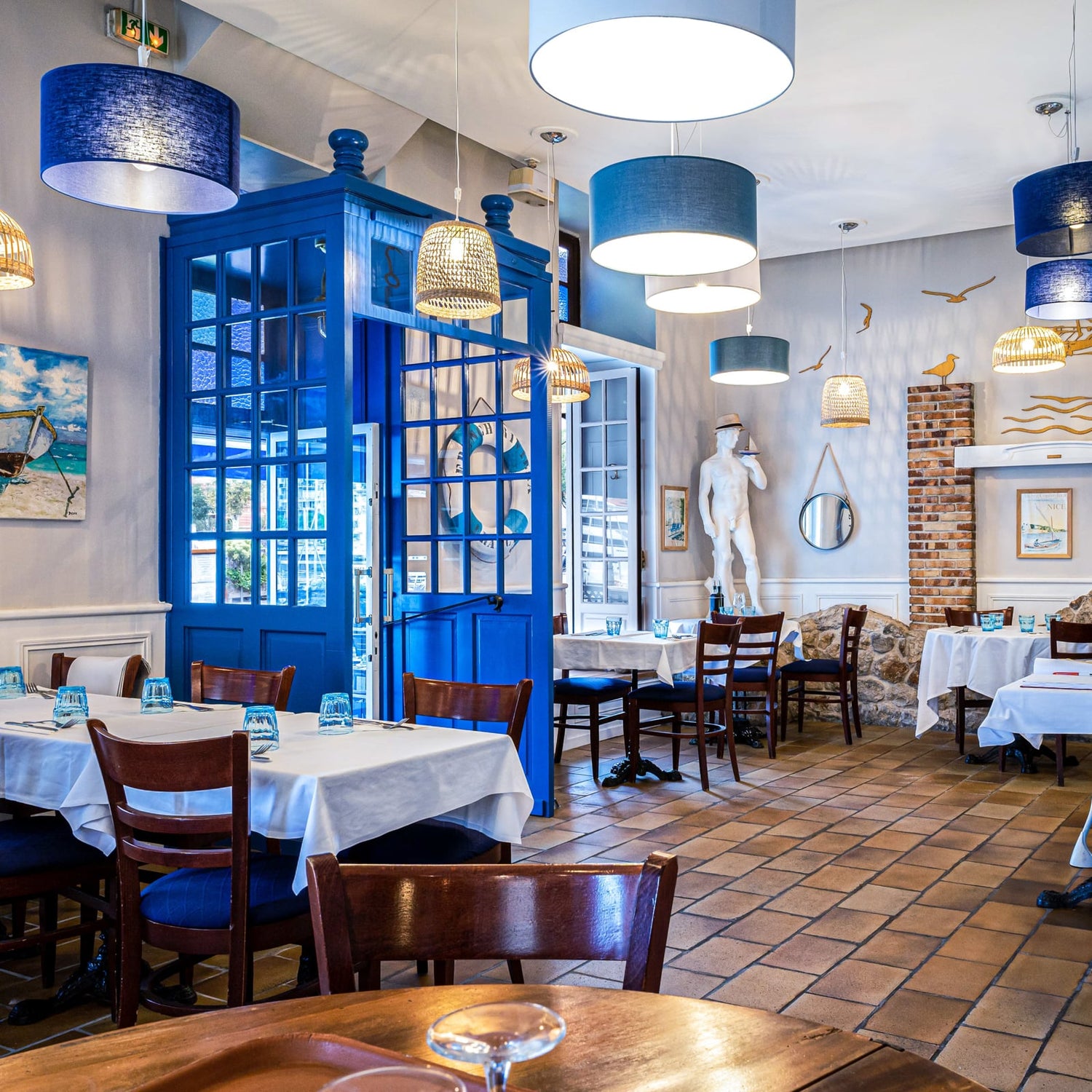 A bright, stylishly decorated restaurant interior with blue pendant lights, a large blue French door, terracotta tile floors, and white tablecloths on dining tables.