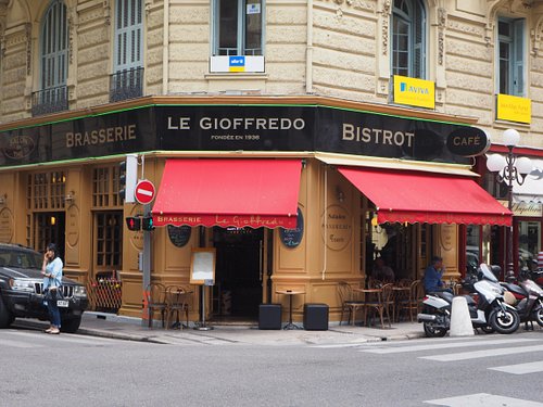 Le Gioffredo Bistrot corner restaurant on a beige stone building, featuring a black signboard with gold lettering, red awnings, outdoor seating, and parked motorcycles on the street.