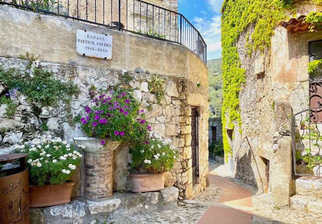 A scenic stone alleyway in a historic Èze Village with beige walls, green ivy, potted white and purple flowers, and a blue sky in the background.
