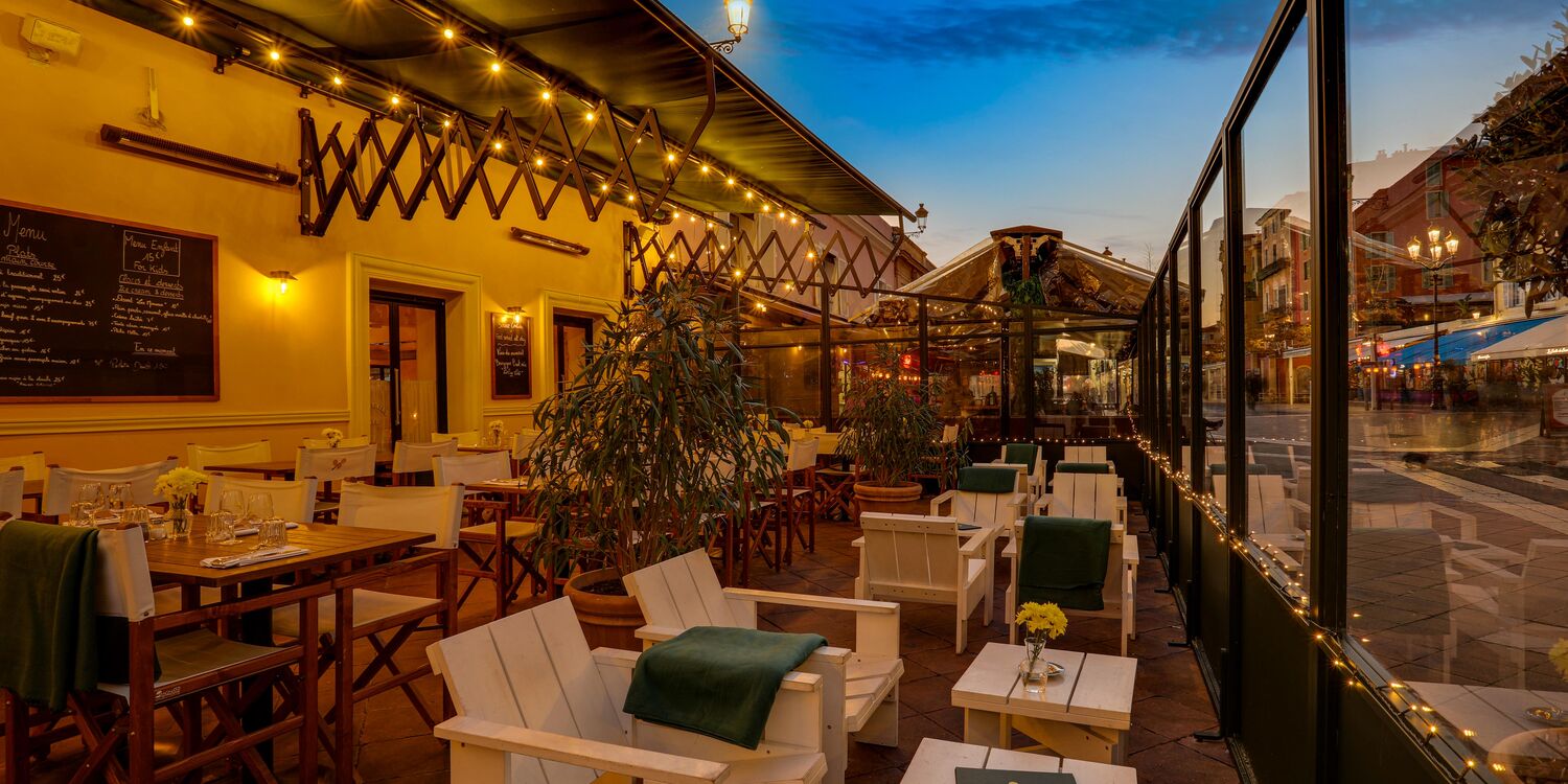 An outdoor patio seating area at dusk with white lounge chairs, small tables, string lights, and a restaurant interior visible on the left, alongside a glass-walled walkway overlooking a street scene.