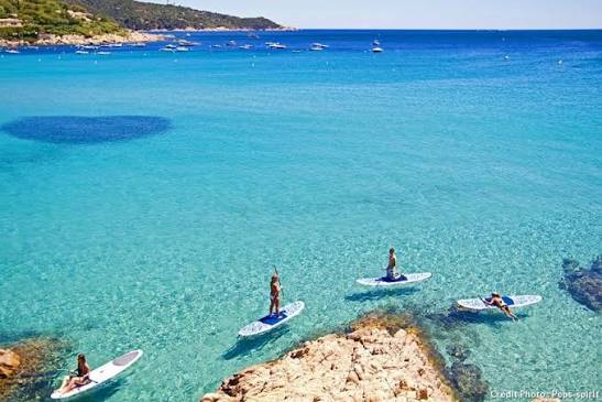 A scenic coastal view of clear turquoise water with several people paddleboarding on the beach, with rocky shoreline and boats in the background.