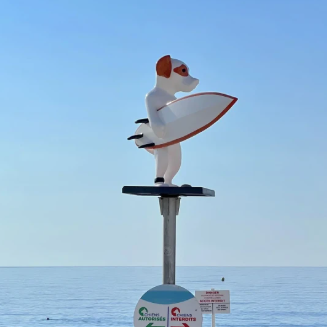 A coastal metal signpost at the beach featuring a white surfboard-shaped bird figure with orange accents, above directional signs, set against a rocky shoreline and blue sea under a clear sky.