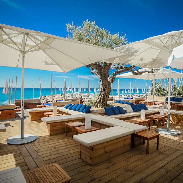Outdoor beachside lounge area with wooden deck seating, large white umbrellas, blue cushions, a central tree, and the blue sea and sky in the background.