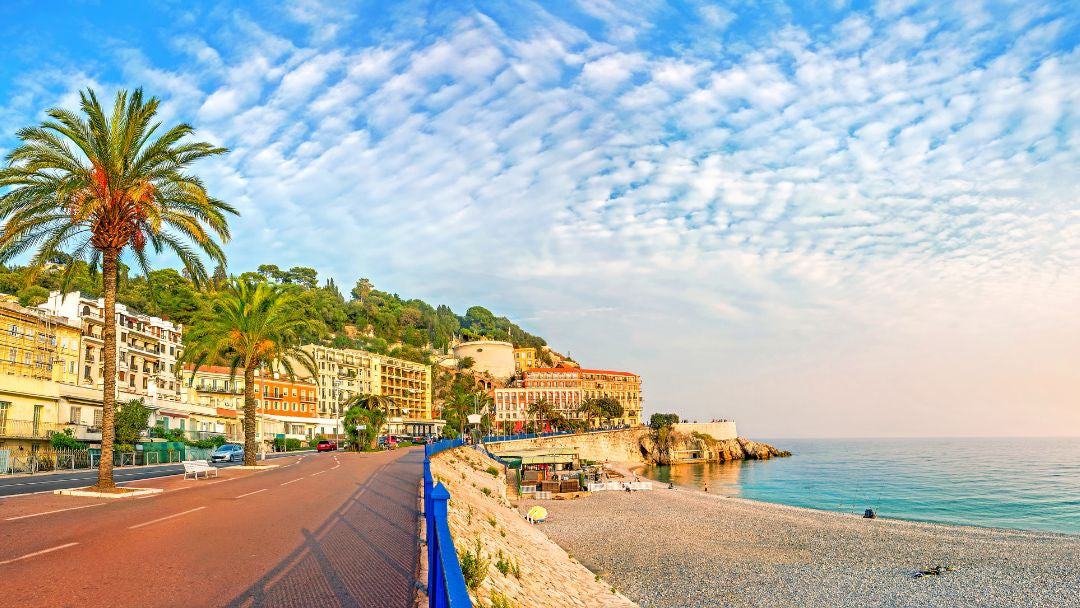 Scenic coastal view with a pebble beach, blue sea, a paved road, palm trees, and buildings on a hillside under a bright sky with scattered clouds.