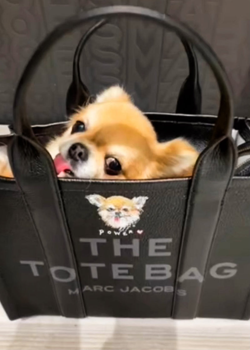 A small tan dog peeking out from a black tote bag with the words 'THE TOTE BAG' and 'MARC JACOBS' printed on the front, with a dog face graphic above.